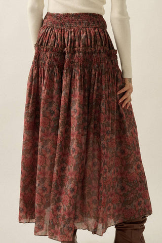 A high-waisted, tiered midi skirt with a smocked waistband and an all-over floral pattern in muted red, brown, and green, styled with a cream ribbed knit top.