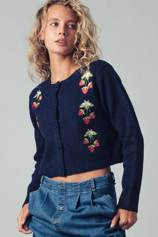 Woman wearing a navy blue cardigan with strawberry embroidery on a plain background