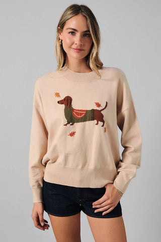A woman modeling a beige crewneck sweater featuring a dachshund graphic with a green sweater, red bow, and orange leaves, paired with dark denim shorts against a solid gray studio background.