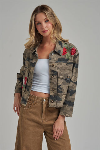 A woman models a cropped camouflage utility jacket featuring red rose embroidery on the chest and sleeves, worn over a white tube top and brown wide-leg jeans against a solid gray studio background.