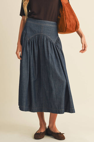 A model wearing a dark blue denim midi skirt with a gathered waist, paired with a dark brown top, brown flats, and carrying a rust-colored suede shoulder bag against a warm beige background.