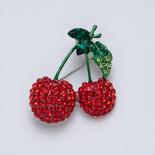 Brooch shaped like a pair of cherries with red gemstones and green leaves on a light gray background