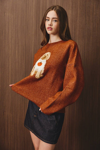 A woman modeling a chunky knit crewneck sweater in a warm rust-orange color, featuring a knitted rabbit motif on the front with a small orange heart accent. She is wearing a dark denim mini skirt against a dark wood-paneled background.