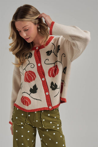 A woman wearing a cream knit cardigan with red trim and red, black, and green pumpkin and vine motifs, paired with olive green polka-dot pants, against a light gray studio background.