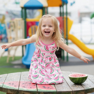 Child in a pink watermelon dress standing on a playground with a watermelon on a table.