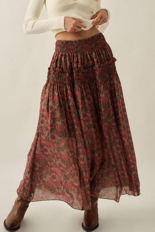 A model wearing a long, flowy maxi skirt with a red and dark floral pattern, paired with a white cropped top and brown ankle boots, against a plain light background.