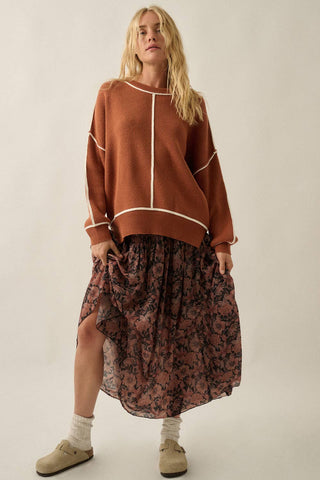 A woman modeling a rust-brown knit sweater with cream contrast stitching and a matching floral-patterned midi skirt, paired with white socks and beige slip-on shoes, against a plain light beige studio background.