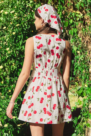Woman wearing a cherry-patterned dress and headscarf standing in front of green foliage.