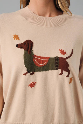 A close-up of a person wearing a cream-colored knit sweater featuring a dachshund wearing a red and green sweater, surrounded by autumn leaves.