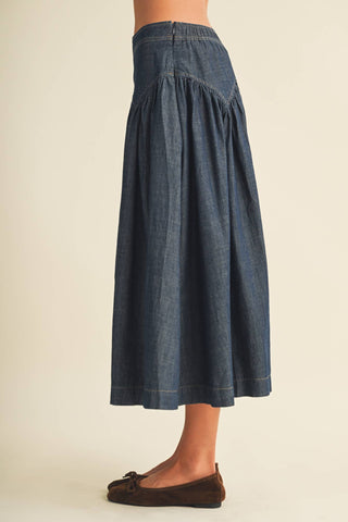 A person wearing a dark blue denim midi skirt with gathered pleats and a smocked waistband, paired with dark brown flat shoes, shown from the waist down against a plain beige background.