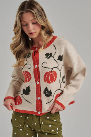 A woman wearing a cream knit cardigan with red trim and black pumpkin vine embroidery, paired with olive green polka-dot pants, against a solid gray studio background.