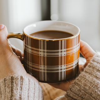 A close-up of a person holding a ceramic coffee mug with a brown, white, and beige plaid pattern and gold trim. The mug is filled with a hot coffee drink, and the person is wearing a cozy beige knit sweater.