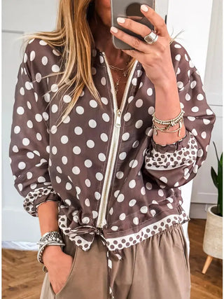 A woman takes a mirror selfie wearing a brown and white polka dot zip-up jacket with a tie detail at the waist, paired with brown pants. She is wearing multiple silver bracelets and rings.
