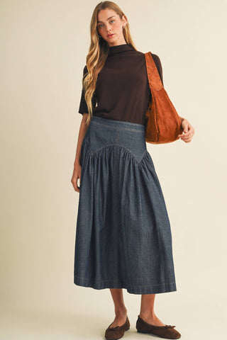 A woman modeling a dark brown mock-neck short-sleeve top and a high-waisted, pleated blue denim midi skirt, carrying a brown suede shoulder bag against a solid beige studio background.