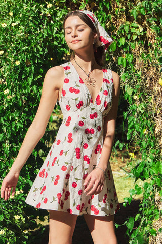 Woman wearing a cherry-patterned dress standing in front of green foliage.