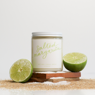 Candle labeled 'Salted Margarita' with two limes on a sandy surface