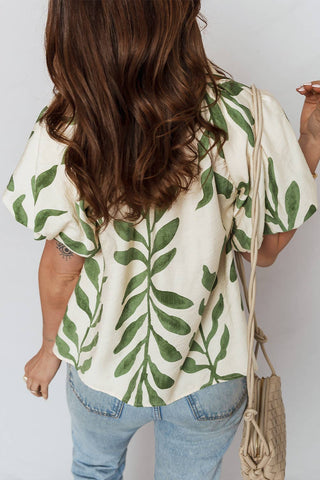 Person wearing a white blouse with green leaf pattern and blue jeans, holding a beige bag.
