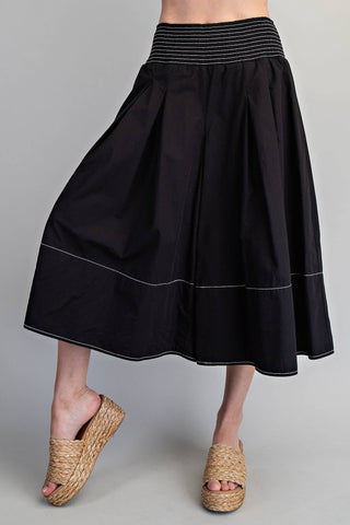 A black pleated midi skirt with white contrast stitching and a striped elastic waistband, shown on a model wearing woven espadrille wedge sandals against a neutral gray background.