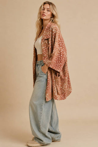 A woman models a rust-colored open-front cardigan with a white speckled pattern, styled over a white crop top and light-wash wide-leg jeans against a solid beige studio background.