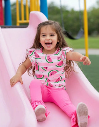 Child sliding down a pink slide at a playground wearing a watermelon print shirt and pink pants.