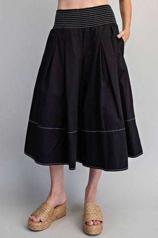 A black pleated midi skirt with a striped elastic waistband and thin white piping details, shown on a model wearing woven platform sandals against a light gray background.