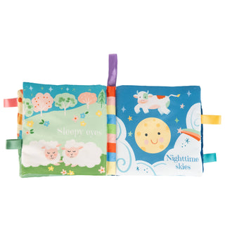 A baby sensory fabric book with two colorful pages, one showing sleeping sheep under a tree on a green background and the other showing a moon and stars on a blue night sky. The pages are held together by a striped spine with a purple tab, and ribbon tabs extend from the sides.