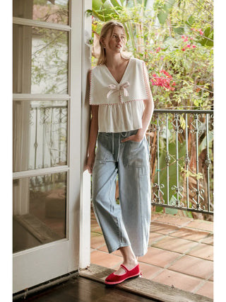 Woman standing on a porch with a garden view, wearing a light-colored top and blue pants.
