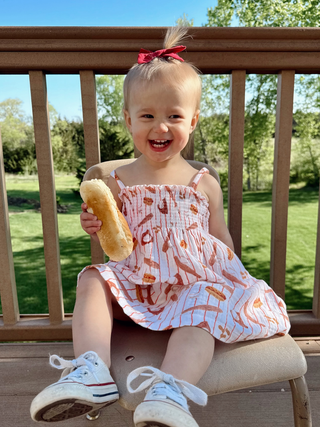 Child sitting on a chair outdoors holding a hot dog, with a wooden deck and green grass in the background.