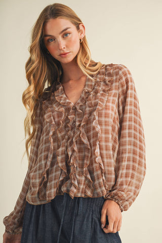 A woman wearing a long-sleeve ruffled blouse with a brown and beige plaid pattern, paired with dark blue jeans, against a plain beige background.