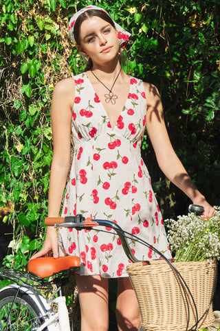 Woman in a cherry-patterned dress standing next to a bicycle with a basket, surrounded by greenery.