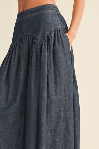 Close-up of a dark blue denim skirt with a gathered waist and side pocket, worn by a model against a light beige background.