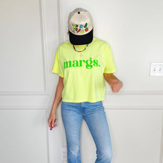 Person wearing a neon yellow 'margs' t-shirt and blue jeans against a white wall.