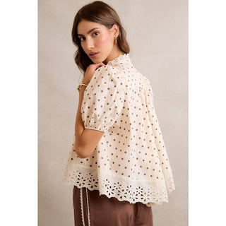 Woman wearing a white blouse with black polka dots and brown pants against a neutral background