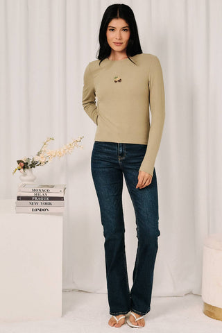 A woman modeling a beige long-sleeve crewneck top with a small embroidered chest detail, paired with dark blue jeans, standing in front of a white curtain backdrop.
