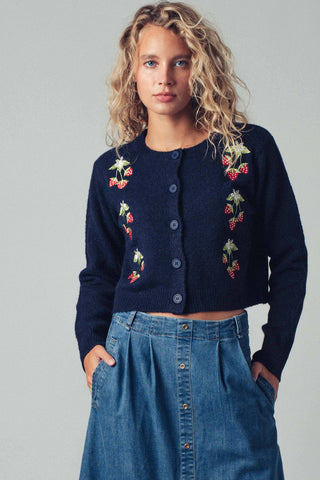 Woman wearing a navy blue cardigan with floral embroidery and a denim skirt on a plain background