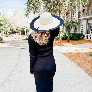 Woman in a black dress and white hat walking on a sidewalk with trees and a building in the background.