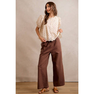 Woman wearing a white blouse with short sleeves and brown wide-leg pants against a beige wall.
