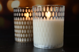 Two lit candles in decorative glass holders on a dark surface with a blurred background.