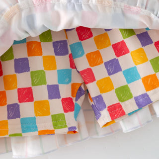 Close-up of a pair of shorts with a colorful checkered pattern made of painted squares in red, orange, yellow, green, blue, and purple on an off-white background. The shorts are layered over a white ruffled skirt or undergarment.