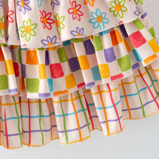 Close-up of a tiered ruffle skirt with a multicolor floral print on the top layer and colorful checkered and plaid patterns on the lower layers, set against a plain light gray background.