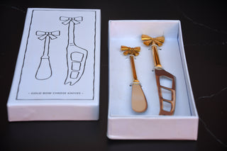 Two gold cheese knives in a white box with packaging illustration on a dark surface.