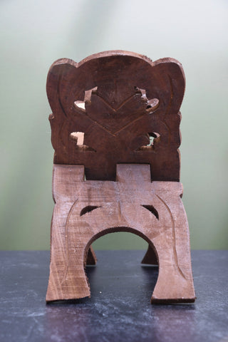 A small carved wooden stool or seat with a decorative cutout backrest and three legs, photographed on a dark tabletop against a soft light green background.