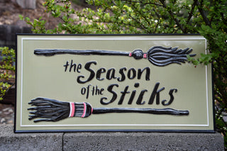 A decorative rectangular sign reading 'the Season of the Sticks' with two raised broomstick illustrations, photographed outdoors against green foliage and a stone wall.