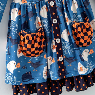 A close-up of a blue Halloween-themed fabric garment, likely a skirt or dress panel, featuring printed ghosts, black cats, crows, spiderwebs, and orange-and-black checkered pockets. The fabric also includes a vertical band with white buttons and orange polka dots, layered over a dark navy hem with orange polka dots.