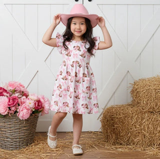 Young girl in a pink floral dress and hat standing on straw with flowers and hay bale in the background.