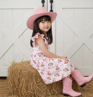 Young girl in a pink dress and cowboy boots sitting on a hay bale with a white wooden background