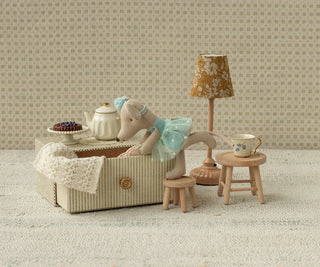 Miniature dollhouse scene with a teddy bear, tea set, and lamp against a patterned wall.