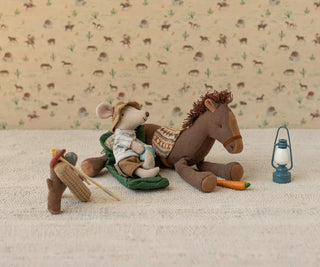 Stuffed mouse in cowboy hat and boots sitting on a horse with a lantern and carrot on a patterned background