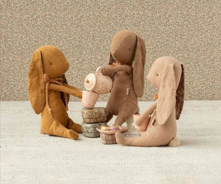Three plush rabbit toys engaged in a tea party on a textured surface with a neutral background.