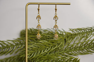 Gold earrings with clear stones on a gold stand with green leaves in the background